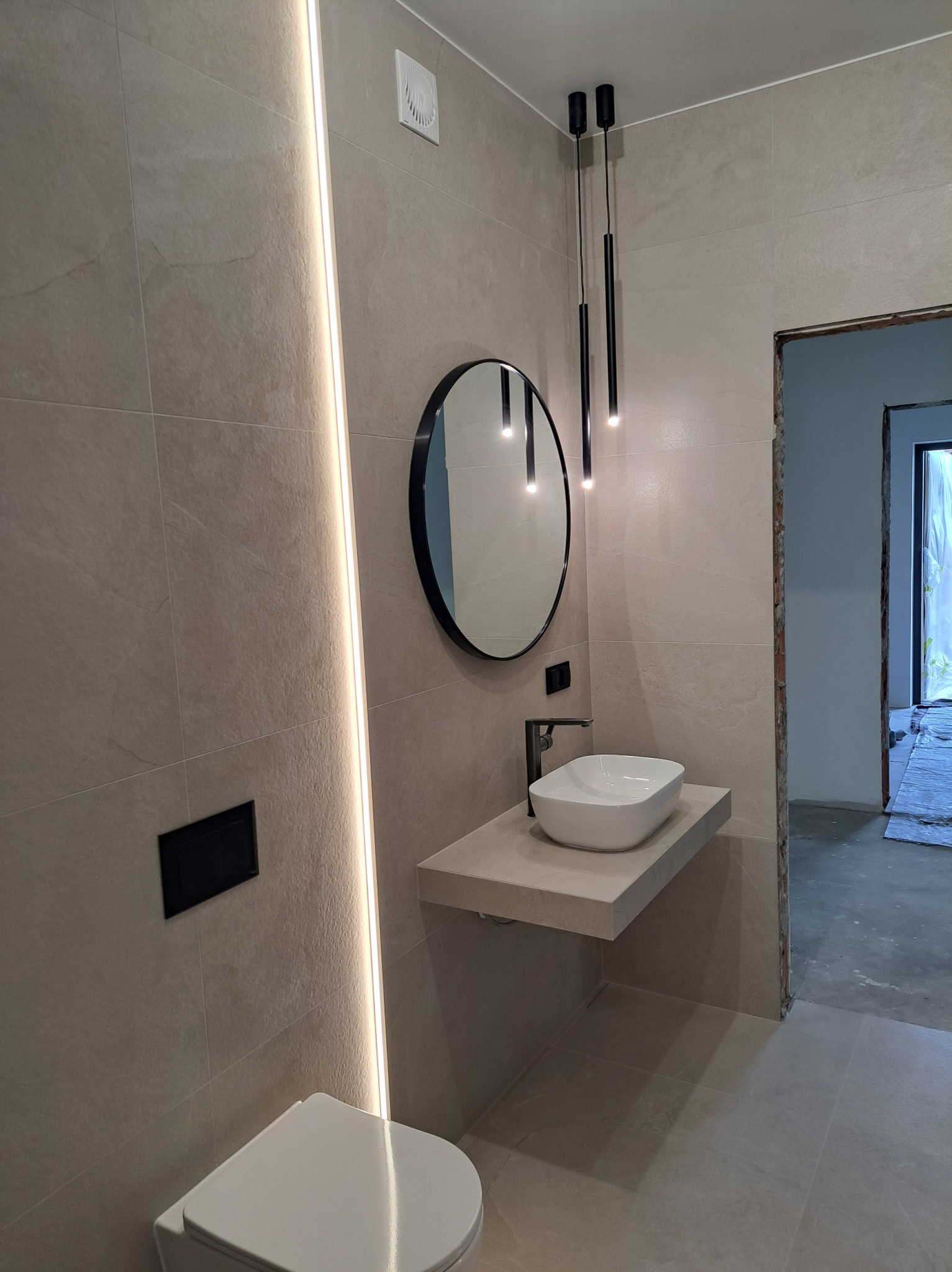 Nowoczesna łazienka z beżowymi płytkami, okrągłym lustrem z czarną ramą, podwieszaną umywalką i minimalistycznym oświetleniem. Widoczny fragment toalety i wejście do innego pomieszczenia.