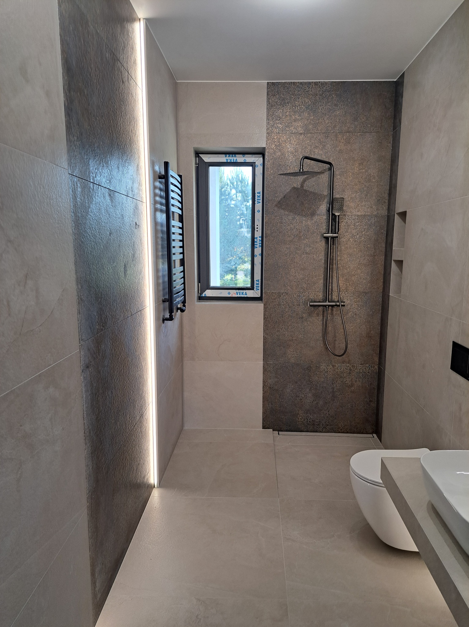 Nowoczesna łazienka z beżowymi płytkami, czarną armaturą i grzejnikiem. Podświetlenie LED wzdłuż ściany. Okno z widokiem na zieleń. Minimalistyczny design.