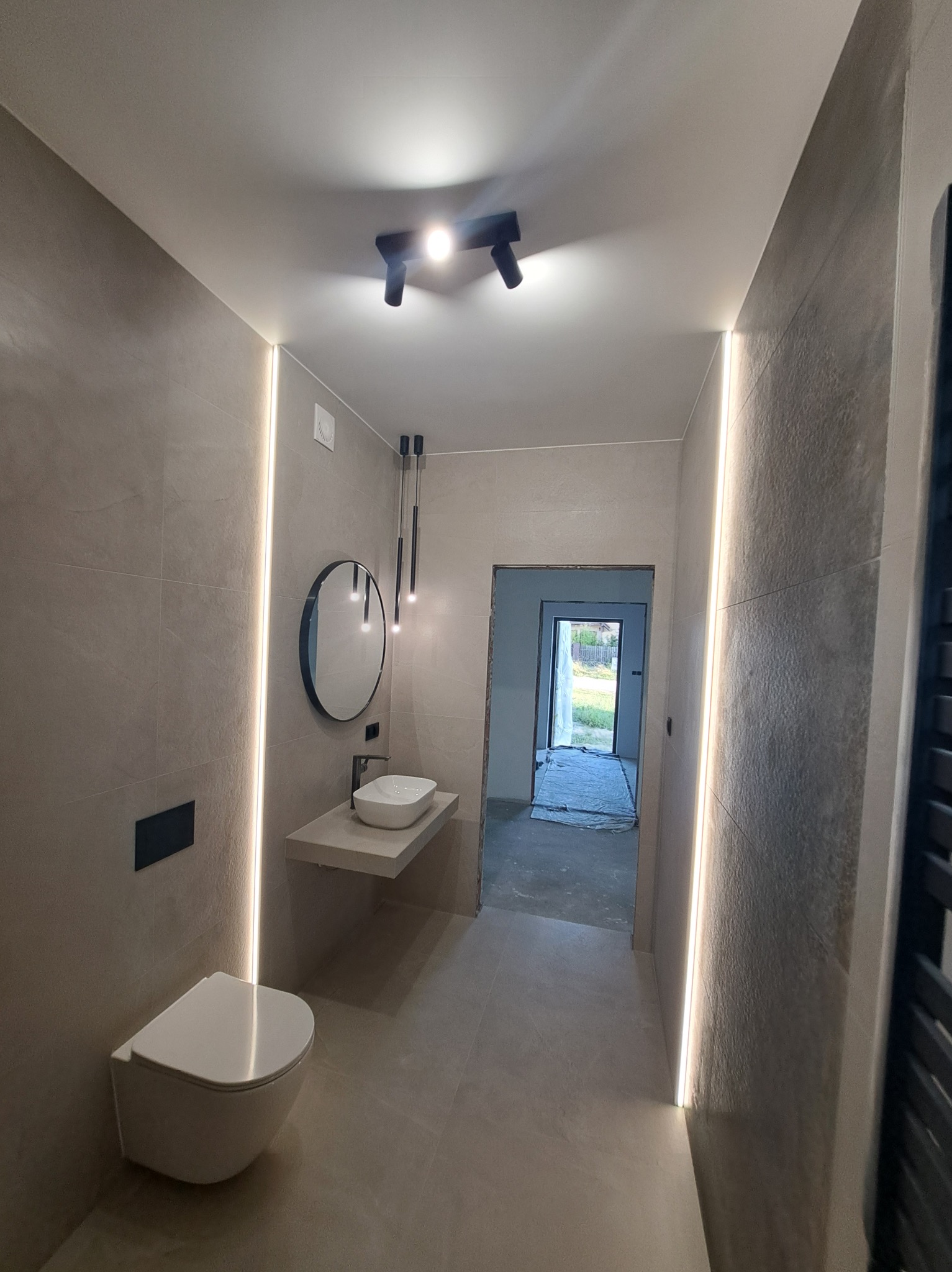 Nowoczesna łazienka w beżowych odcieniach z podświetleniem LED, wiszącą toaletą i okrągłym lustrem nad umywalką. Widok na sąsiednie pomieszczenie.