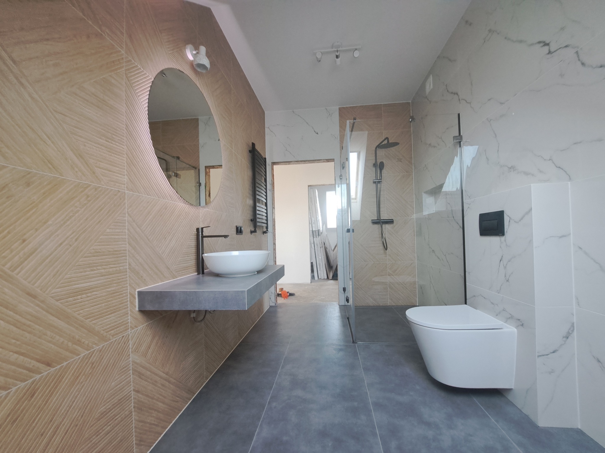 Nowoczesna łazienka z beżowo-białymi płytkami, okrągłym lustrem nad umywalką na szarym blacie i podwieszaną toaletą. Widoczna kabina prysznicowa.