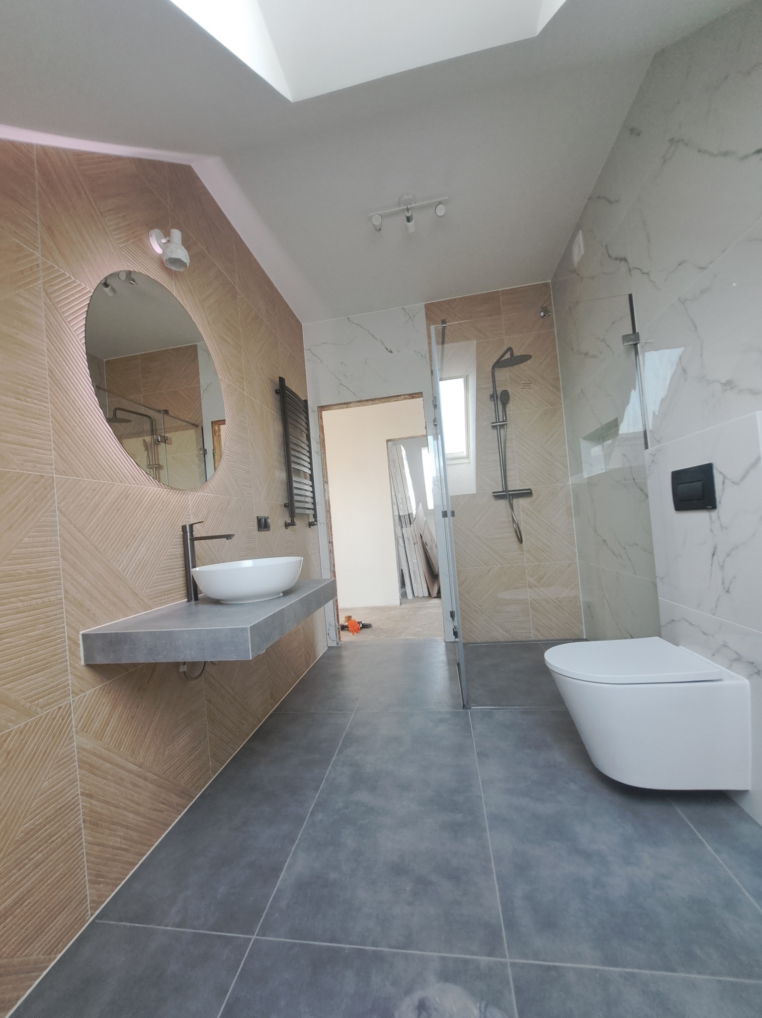 Nowoczesna łazienka z beżowymi i białymi płytkami, szarą podłogą, owalnym lustrem i minimalistyczną armaturą. Widoczny fragment kabiny prysznicowej i toalety.