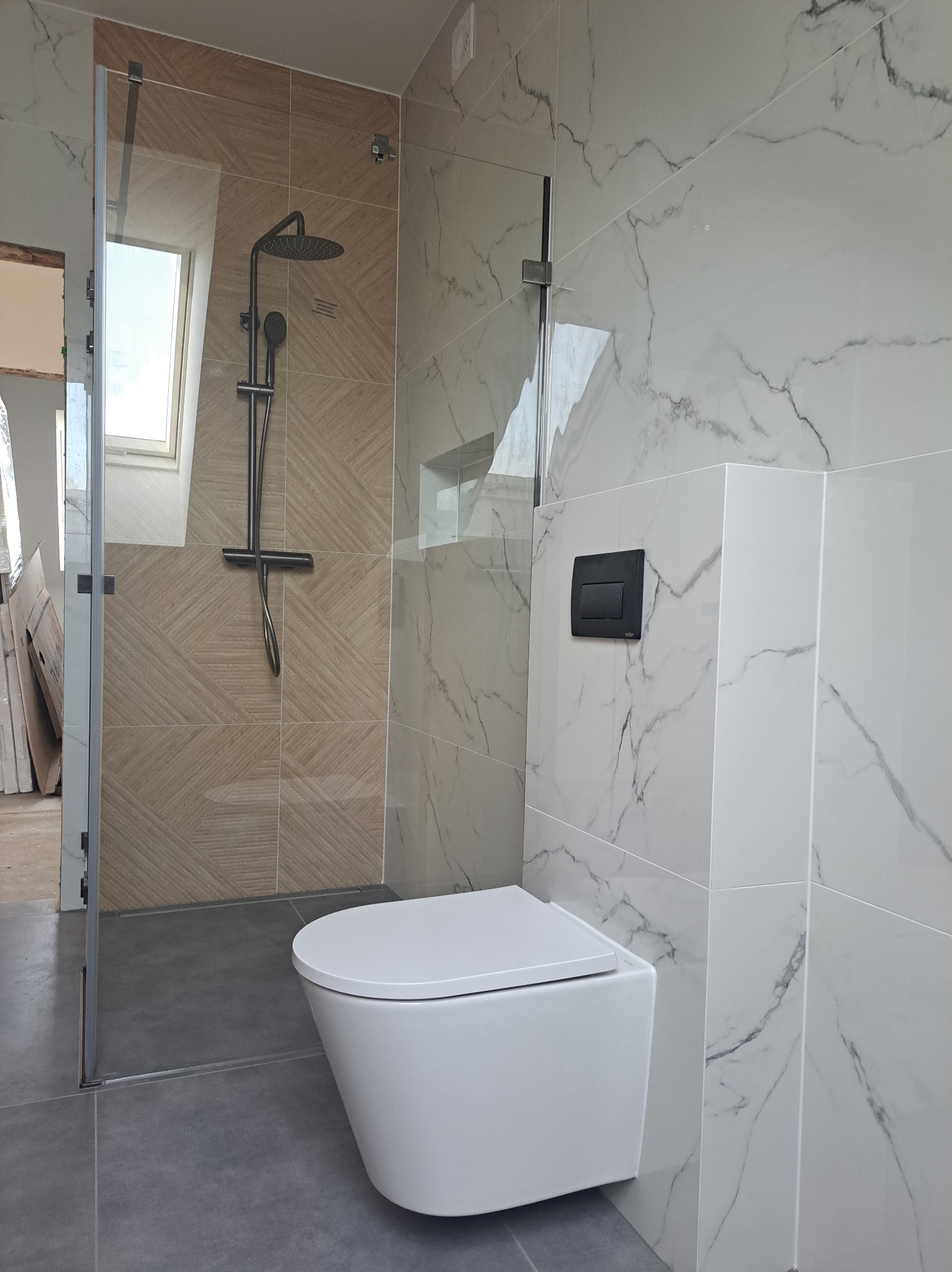 Nowoczesna łazienka z kabiną prysznicową typu walk-in, wiszącą toaletą i marmurowymi płytkami. Uwagę zwraca okno dachowe nad prysznicem.