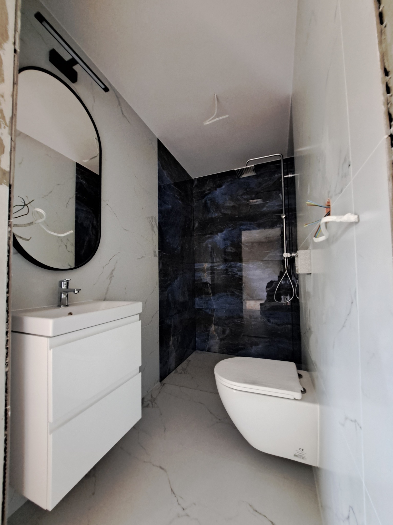 Nowoczesna łazienka z umywalką na szafce, wiszącą toaletą i kabiną prysznicową wyłożoną ciemnym marmurem. Lustro owalne z czarną ramą. Minimalistyczny design.