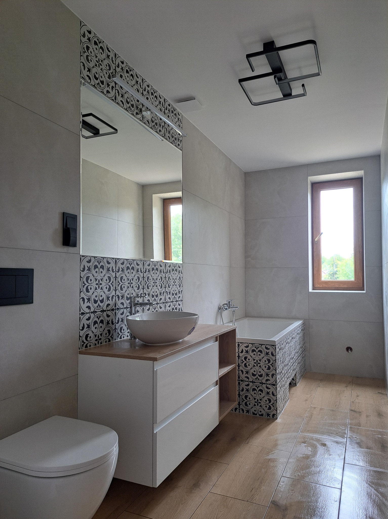 Jasna łazienka z drewnianą podłogą i wzorzystymi płytkami nad wanną i umywalką. Minimalistyczna armatura i oświetlenie.