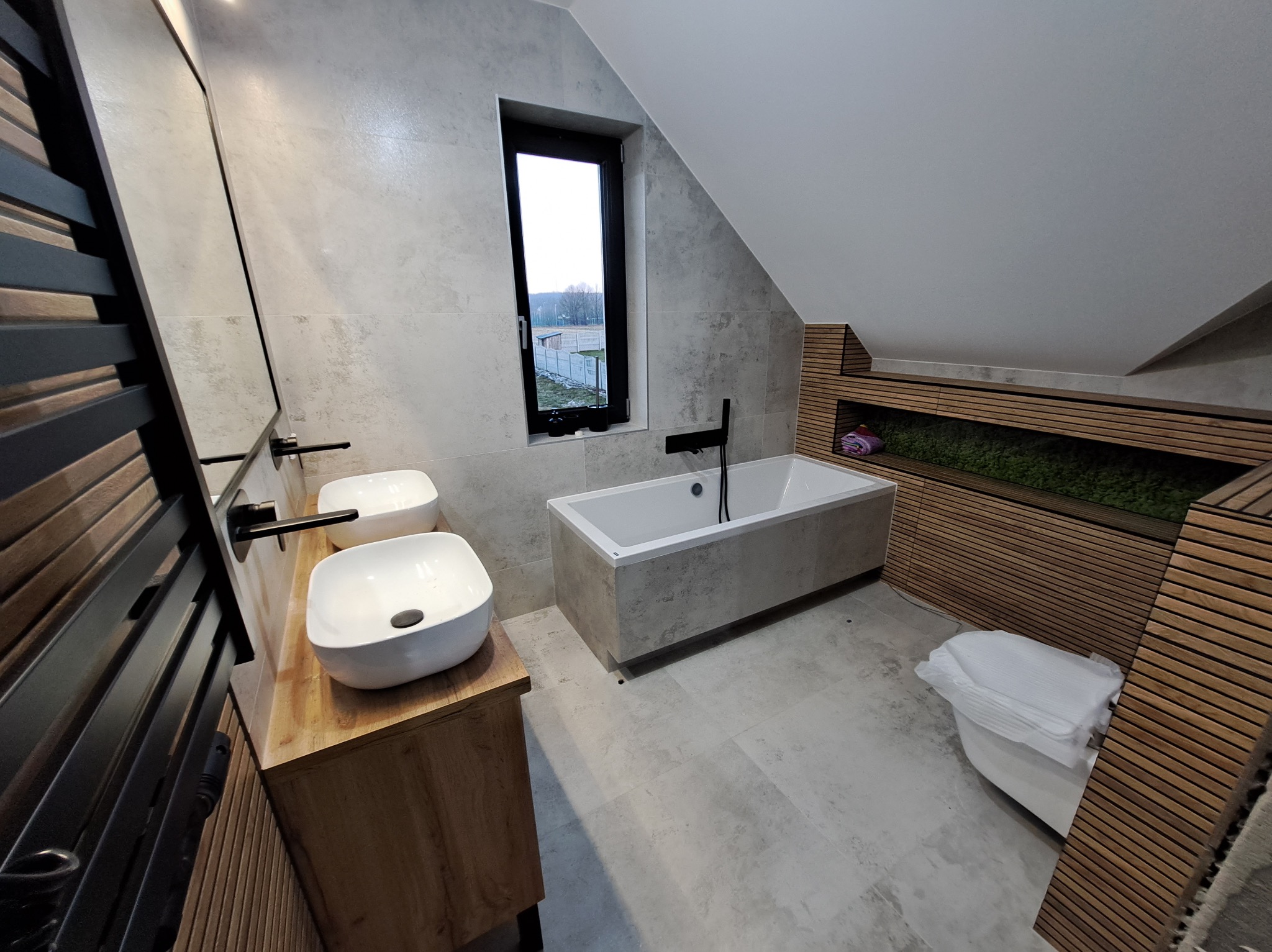 Nowoczesna łazienka na poddaszu z dwoma umywalkami na drewnianym blacie, wanną i oknem. Ściany wyłożone szarymi płytkami, drewniane lamele dekoracyjne.