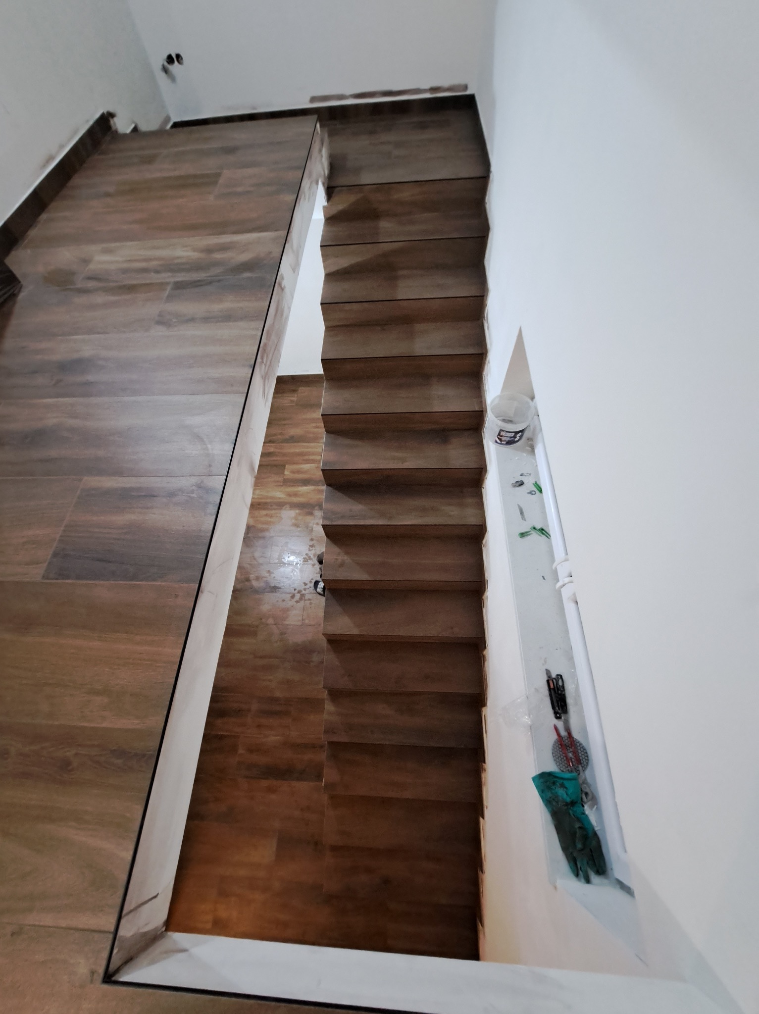 Ukończone schody wyłożone płytkami imitującymi drewno, widok z góry. Na ścianie wiszą rękawice robocze i narzędzia. Podłoga również wyłożona płytkami.