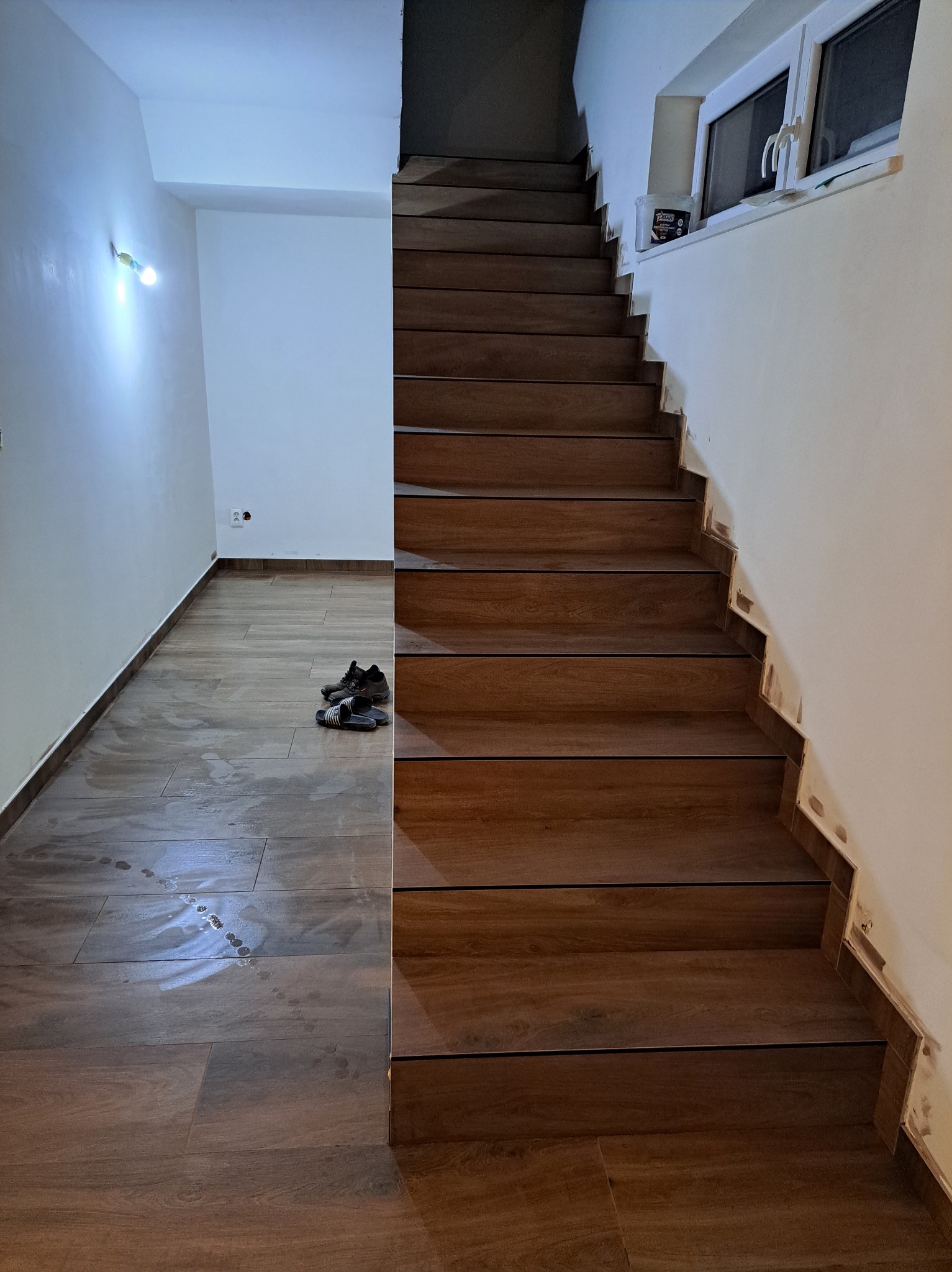 Wykończone schody z drewnopodobnych płytek, prowadzące na górę. Na podłodze widoczne ślady po umyciu i para butów. Ściany pomalowane na biało.