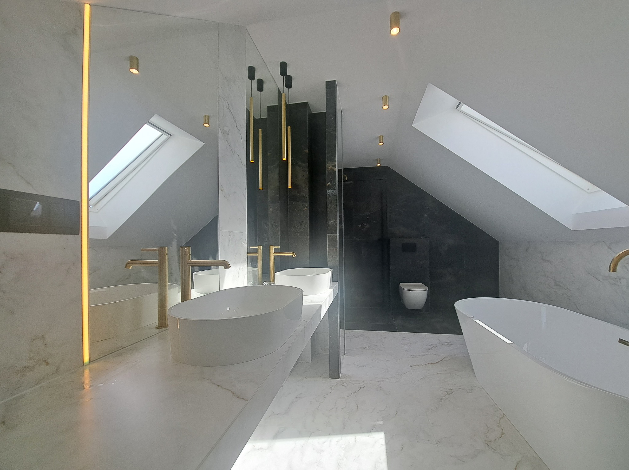Nowoczesna łazienka na poddaszu z marmurowymi płytkami, owalnymi umywalkami, złotą armaturą i oświetleniem punktowym. Widoczne okno dachowe i wolnostojąca wanna.