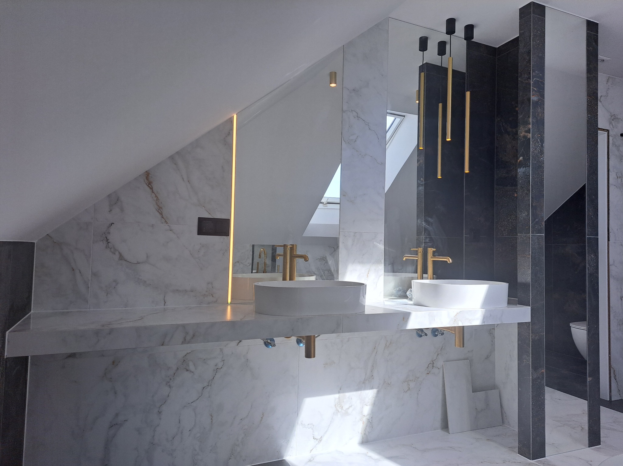 Nowoczesna łazienka na poddaszu z marmurowymi płytkami, złotą armaturą i wiszącymi lampami. Umywalki nablatowe, lustro z podświetleniem LED.