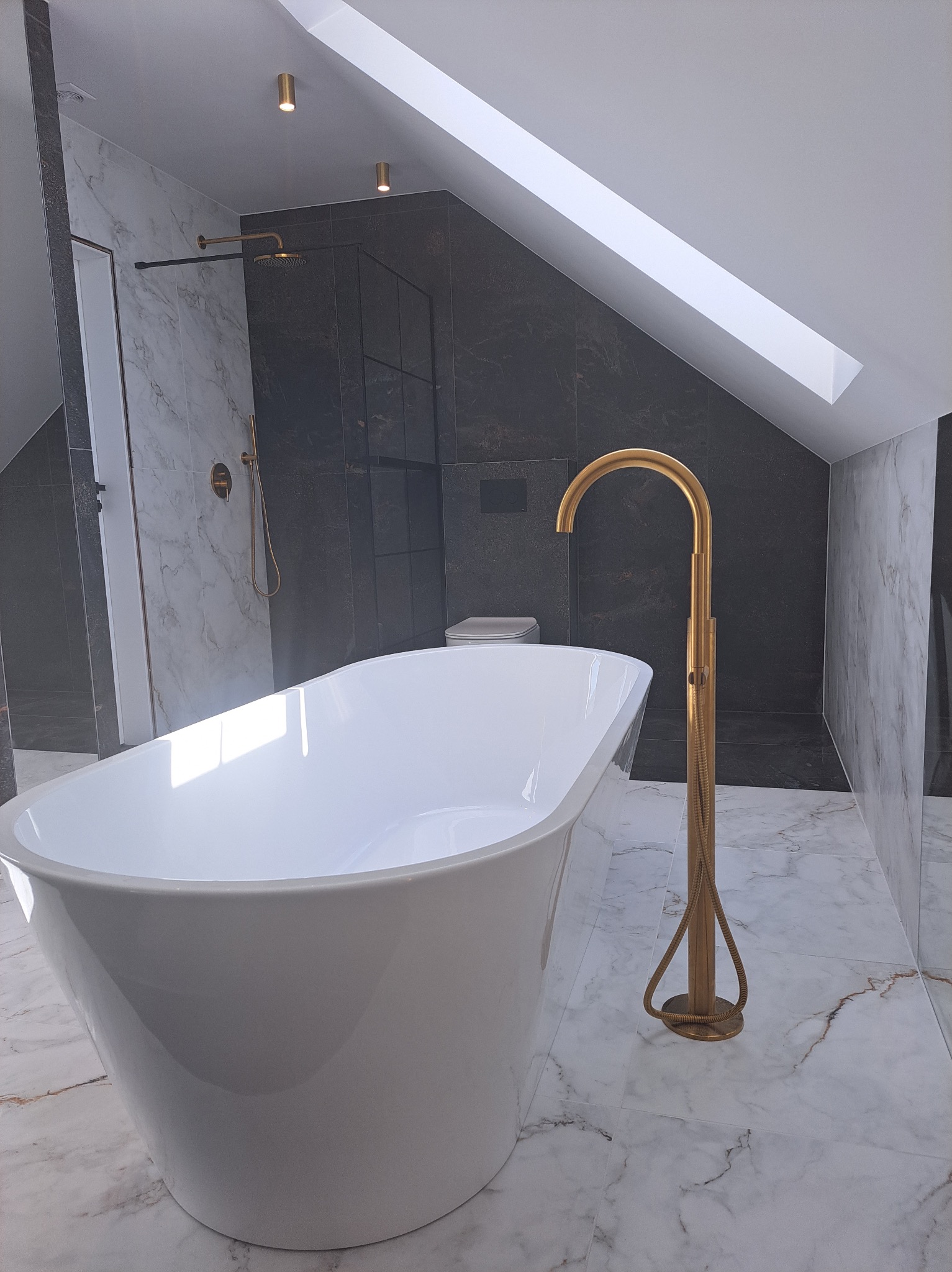 Nowoczesna łazienka na poddaszu z wolnostojącą białą wanną i złotą armaturą na tle marmurowych płytek i kabiny prysznicowej.