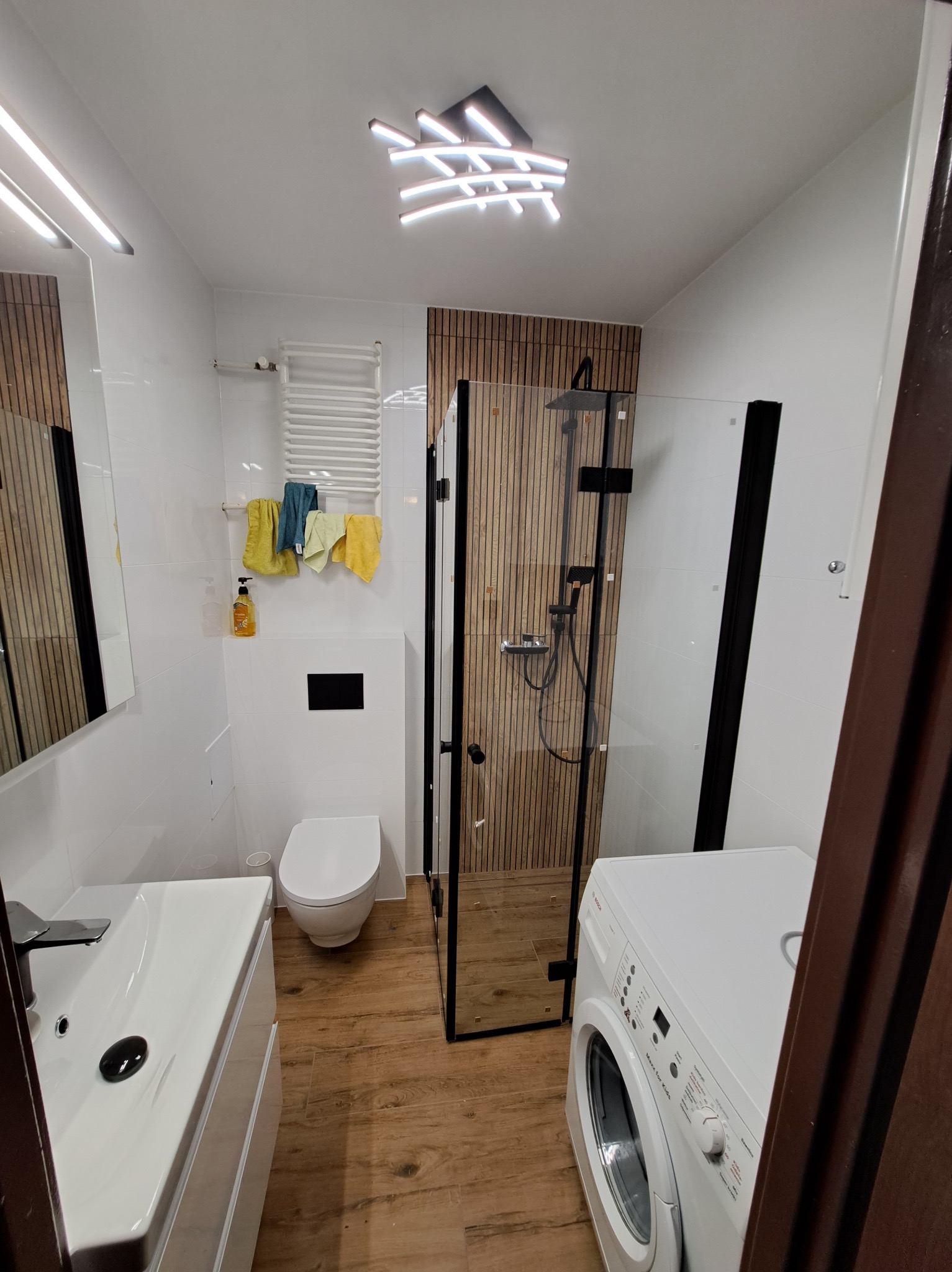 Nowoczesna łazienka z kabiną prysznicową typu walk-in, drewnianymi panelami i białymi płytkami. Umywalka z szafką, toaleta i pralka tworzą funkcjonalną przestrzeń.