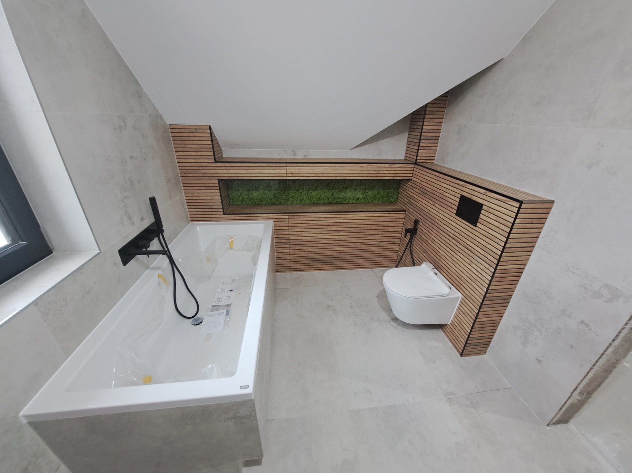 Nowoczesna łazienka na poddaszu z wanną w folii, wiszącą toaletą i drewnianą zabudową z zielonym akcentem. Szare płytki na ścianach i podłodze.