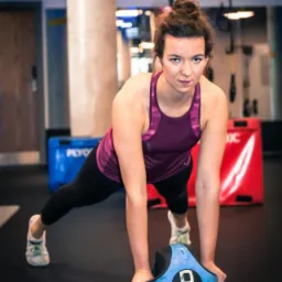 Kobieta w sportowym stroju wykonuje pompkę z użyciem niebieskiej piłki obciążeniowej w siłowni, patrząc w obiektyw.