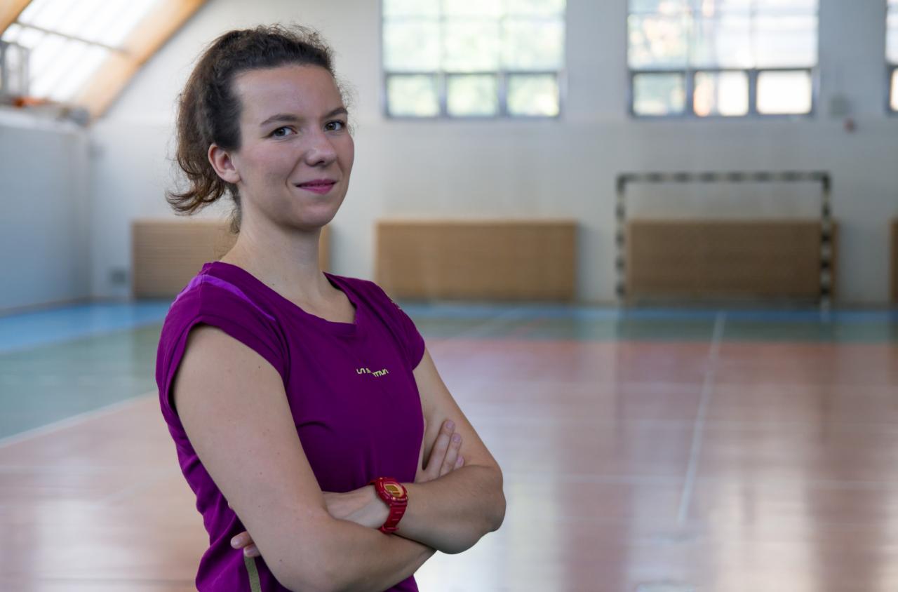 Uśmiechnięta kobieta w fioletowej koszulce sportowej, z założonymi rękami i czerwonym zegarkiem, stojąca w hali sportowej z bramkami w tle.