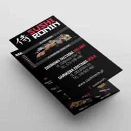 Czarna ulotka reklamowa Sushi Ronin z zestawami sushi, adresami dostawy w Bielanach i Woli oraz danymi kontaktowymi, leżąca na jasnym tle pod kątem.