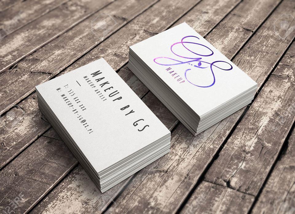 Dwa stosy wizytówek na drewnianym tle. Jedna wizytówka z napisem 'MAKEUP BY GS MAKEUP ARTIST', a druga z logo 'GS MAKEUP' w kolorze fioletowo-różowym.