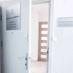 Tabliczka informacyjna na drzwiach wejściowych do kancelarii z napisem 'Kancelaria Opolska', widoczny fragment wnętrza z drzwiami i elementami wyposażenia.