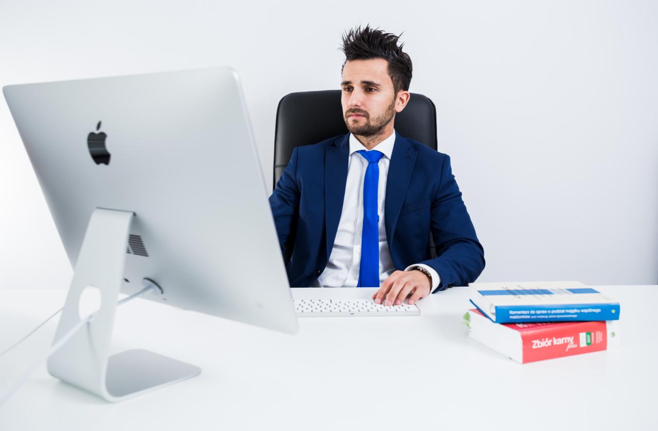 Mężczyzna w niebieskim garniturze i krawacie siedzi przy biurku z komputerem iMac i książkami o tematyce prawnej.