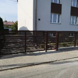 Nowoczesna, pozioma, brązowa konstrukcja ogrodzeniowa przed dwukondygnacyjnym domem z szarą elewacją i brązowymi akcentami, widoczne kostki brukowe chodnika.