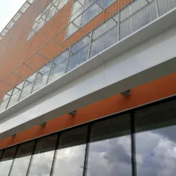 Pomarańczowa fasada budynku biurowego z metalową siatką i dużymi oknami, w których odbijają się chmury.