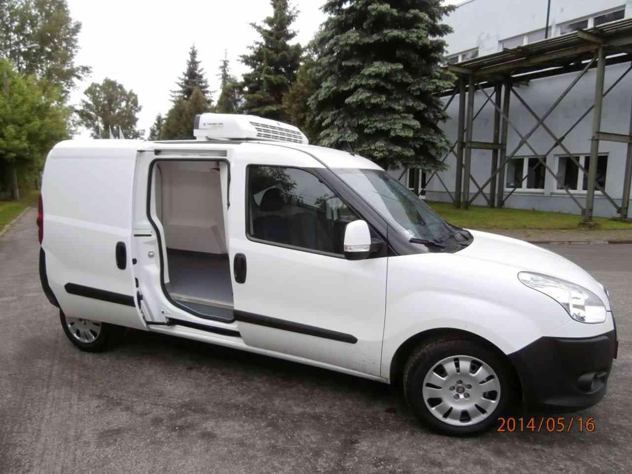 Biały furgon chłodniczy z otwartymi drzwiami bocznymi, widoczny agregat chłodniczy na dachu, zaparkowany na zewnątrz budynku.