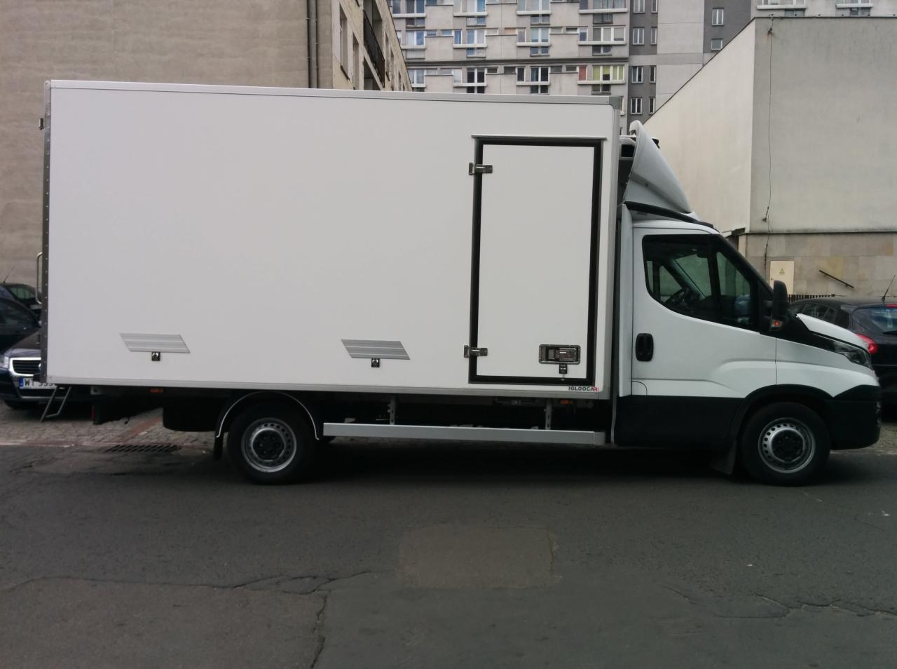 Biały samochód dostawczy z bocznymi drzwiami, zaparkowany na ulicy w miejskiej zabudowie.