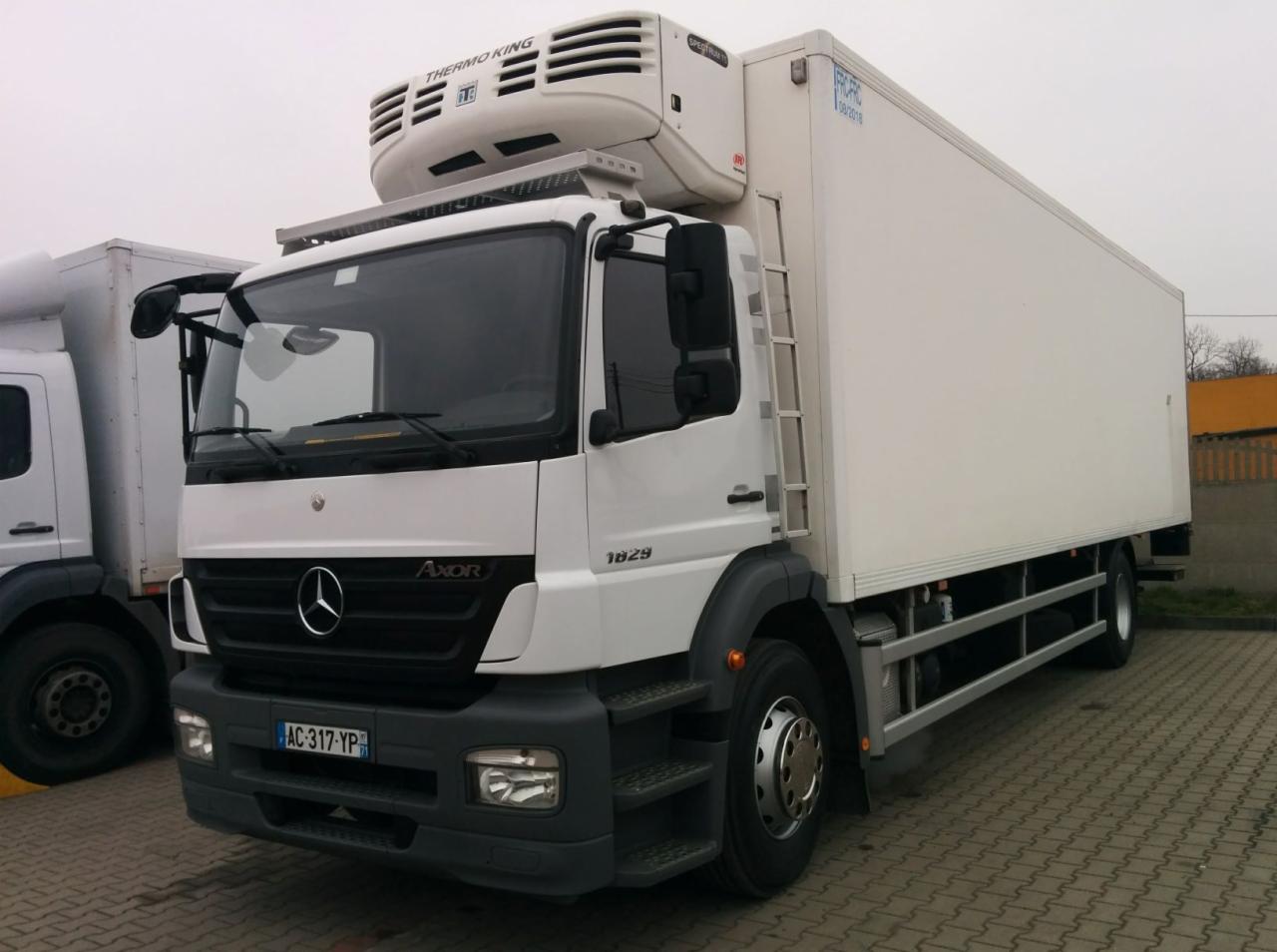 Biała ciężarówka chłodnia marki Mercedes Axor 1829 z agregatem Thermo King, zaparkowana na kostce brukowej w pochmurny dzień.