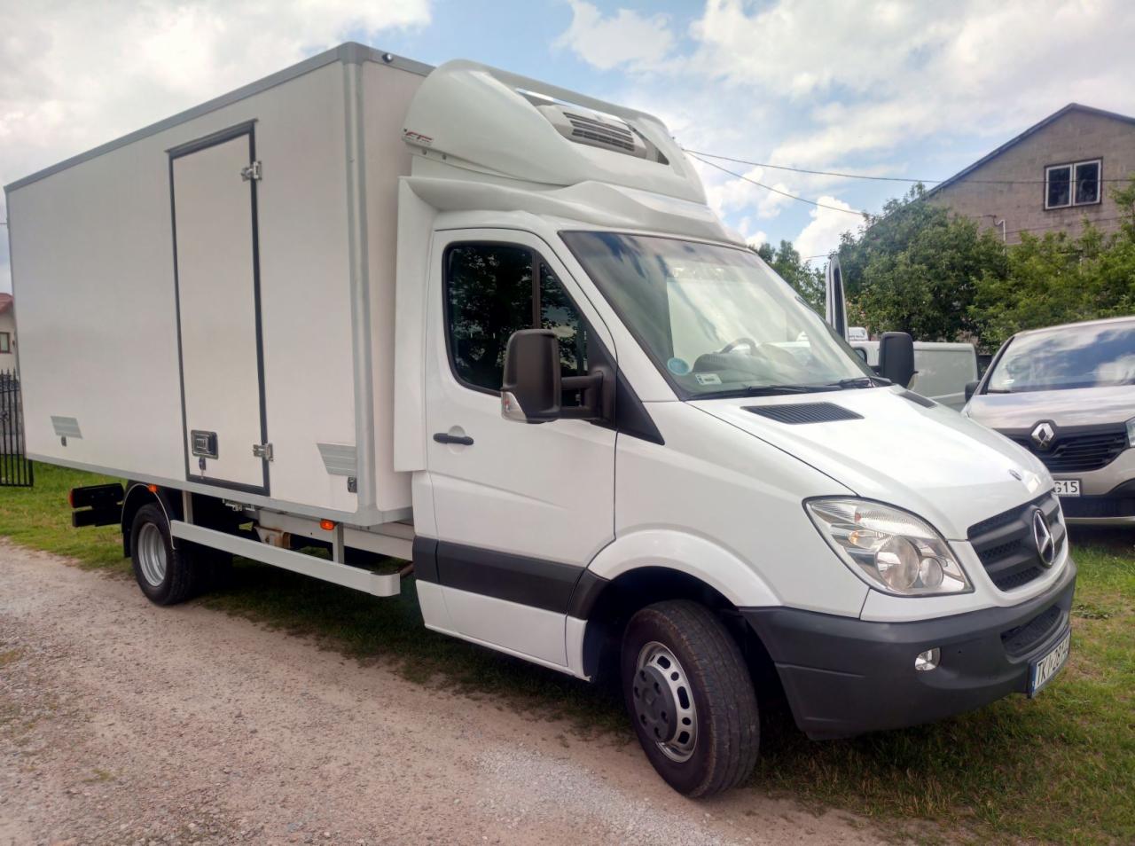 Biały samochód dostawczy typu furgon chłodnia marki Mercedes-Benz Sprinter, zaparkowany na żwirowej drodze, z domem w tle.