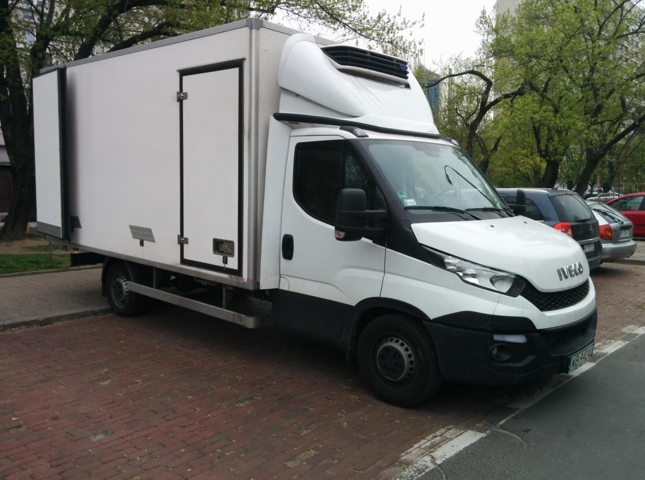 Biały samochód dostawczy Iveco z chłodnią, zaparkowany na chodniku z czerwonej kostki, widok z boku na tle drzew i innych samochodów.
