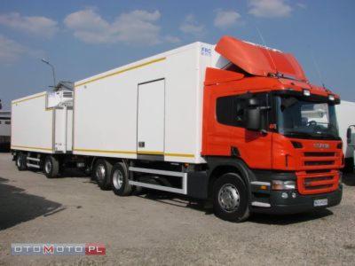 Pomarańczowo-biała ciężarówka Scania z chłodnią i przyczepą na parkingu w słoneczny dzień.