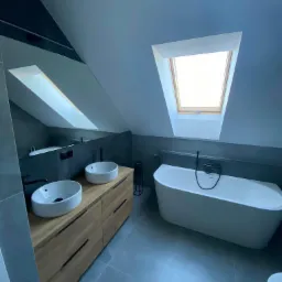 Nowoczesna łazienka na poddaszu z dwoma umywalkami nablatowymi na drewnianej szafce, wolnostojącą wanną i oknem dachowym.