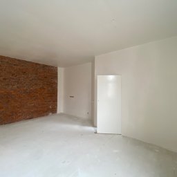 USŁUGI BUDOWLANE GRZEGORZ DZIERGAS - Wnętrze pokoju w trakcie prac wykończeniowych: ściany częściowo pomalowane na biało, fragment ściany z czerwoną cegłą, podłoga pokryta warstwą wyrównującą, widoczne gniazdka elektryczne.