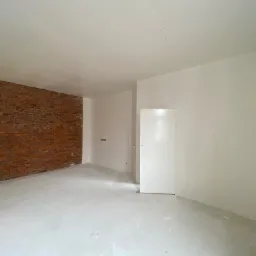 Wnętrze pokoju w trakcie prac wykończeniowych: ściany częściowo pomalowane na biało, fragment ściany z czerwoną cegłą, podłoga pokryta warstwą wyrównującą, widoczne gniazdka elektryczne.