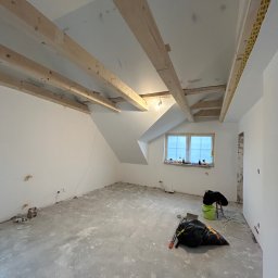 USŁUGI BUDOWLANE GRZEGORZ DZIERGAS - Pokój na poddaszu w trakcie prac wykończeniowych; widoczne odsłonięte drewniane belki stropowe, surowy beton na podłodze z resztkami zaprawy, worki ze śmieciami, nieotynkowane fragmenty ścian...