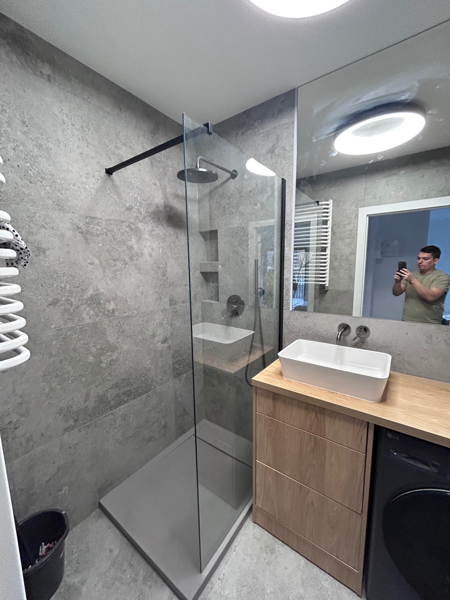 Nowoczesna łazienka z kabiną prysznicową typu walk-in, szare płytki imitujące beton, biała umywalka nablatowa na drewnianej szafce i odbicie osoby robiącej zdjęcie w lustrze.