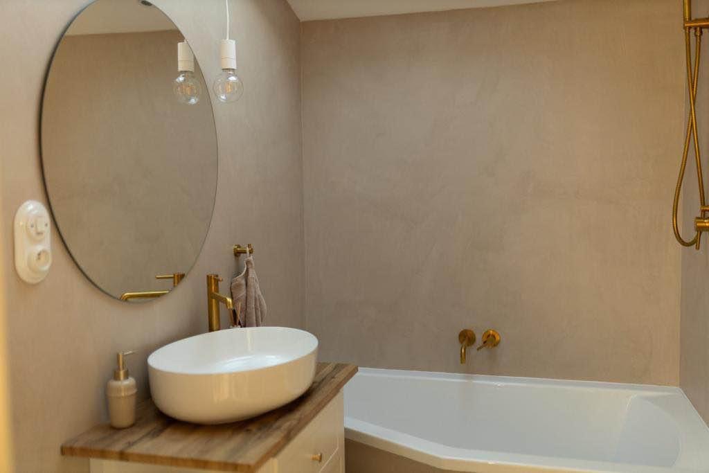 Wykończona łazienka w beżowych odcieniach z owalnym lustrem, umywalką nablatową na drewnianym blacie, złotą armaturą i gładzią na ścianach.