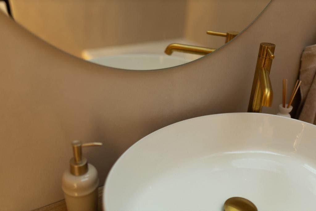 Biała umywalka nablatowa z złotą armaturą i dozownikiem mydła, odbicie w lustrze o nieregularnym kształcie.