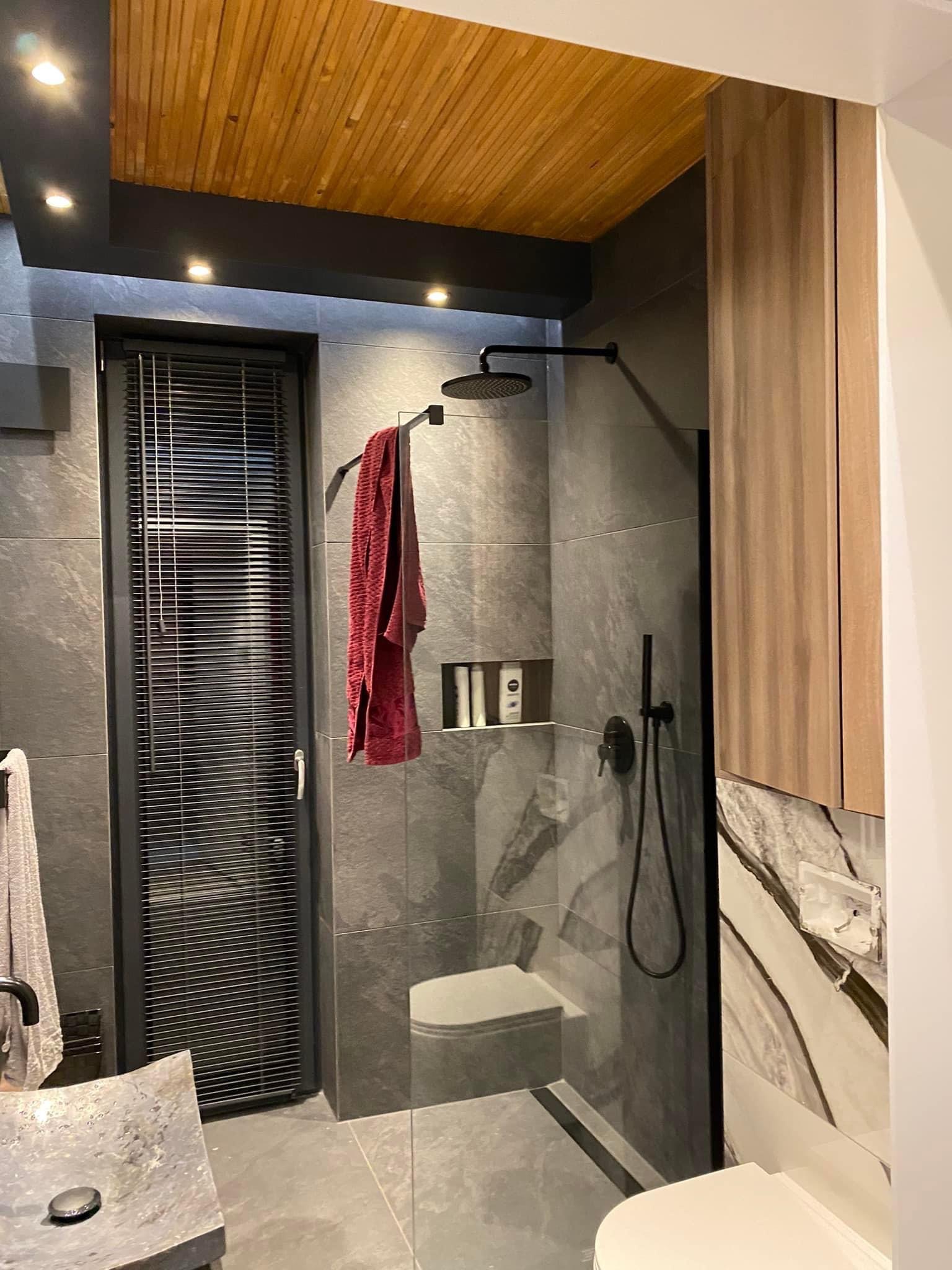 Nowoczesna łazienka z szarymi płytkami imitującymi kamień, drewnianym sufitem i kabiną prysznicową typu walk-in z czarną armaturą, widok na umywalkę z szarego kamienia i drewnianą szafkę.