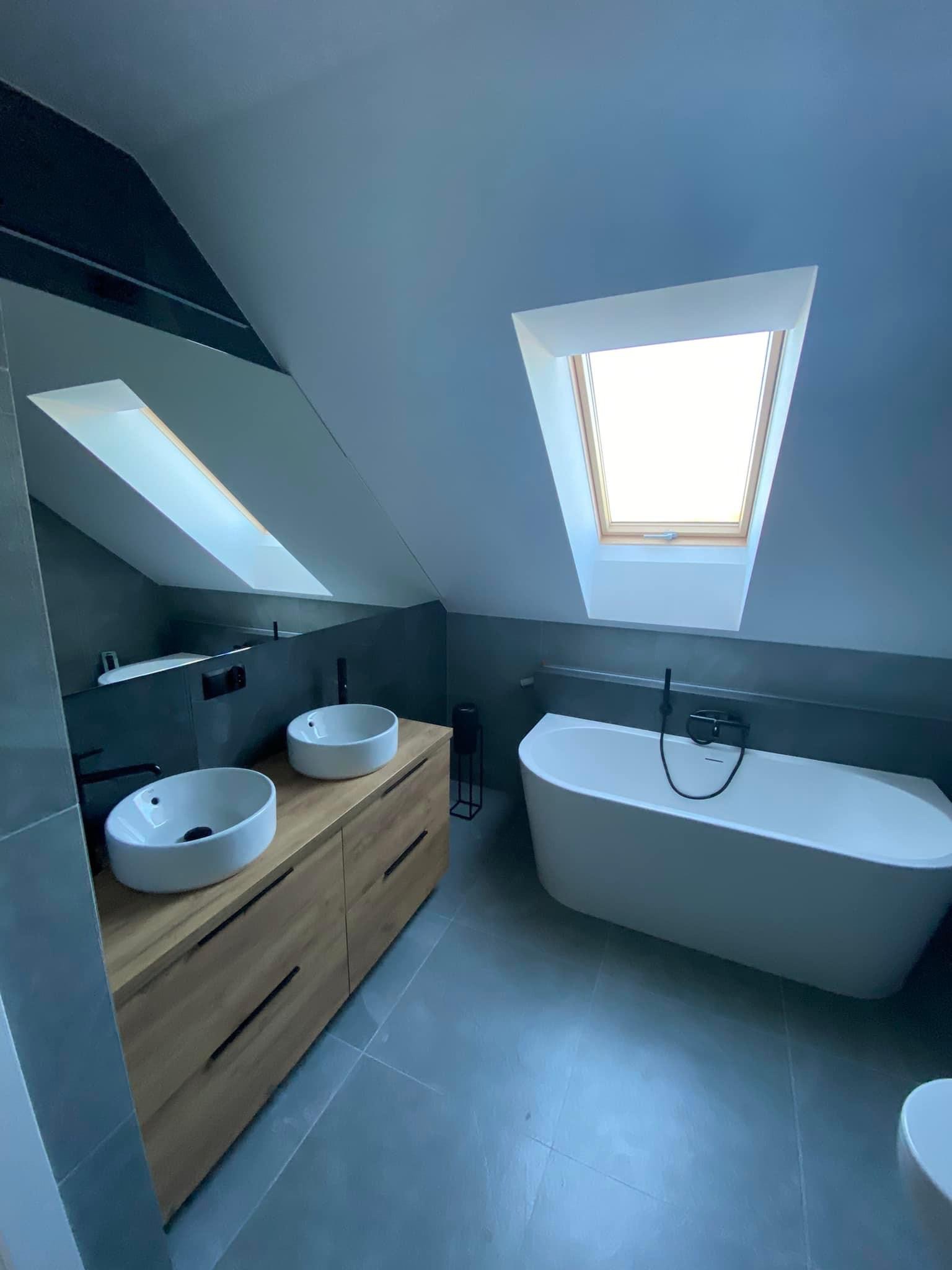 Nowoczesna łazienka na poddaszu z dwoma umywalkami nablatowymi na drewnianej szafce, wolnostojącą wanną i oknem dachowym.
