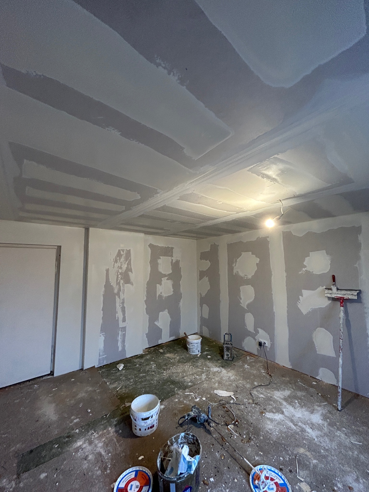 Wnętrze w trakcie prac przygotowawczych do malowania; ściany i sufit pokryte fragmentarycznie gładzią, widoczne narzędzia i akcesoria malarskie, podłoga zabezpieczona folią, w tle wisząca żarówka.