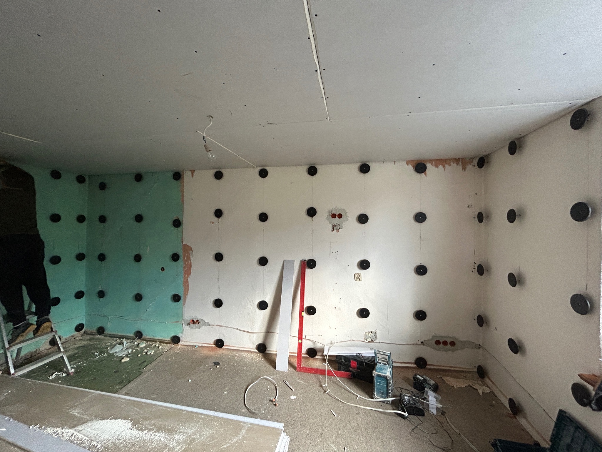 Montaż płyty karton gips na ścianach z użyciem kołków montażowych, widoczny fragment ściany z zieloną płytą, poziomica, szlifierka i drabina.