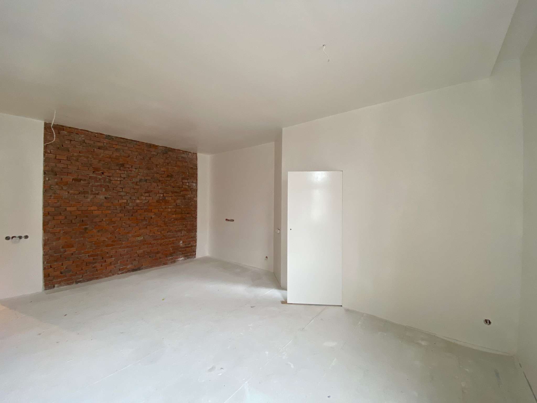 Wnętrze pokoju w trakcie prac wykończeniowych: ściany częściowo pomalowane na biało, fragment ściany z czerwoną cegłą, podłoga pokryta warstwą wyrównującą, widoczne gniazdka elektryczne.