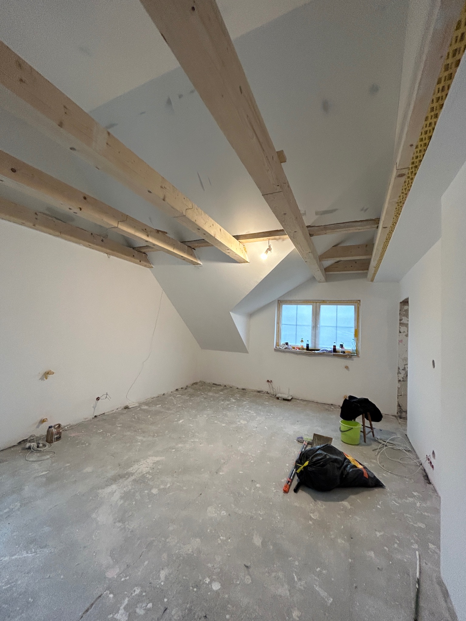 Pokój na poddaszu w trakcie prac wykończeniowych; widoczne odsłonięte drewniane belki stropowe, surowy beton na podłodze z resztkami zaprawy, worki ze śmieciami, nieotynkowane fragmenty ścian...