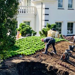 MEGIDO_Serwis - Układanie darni na wzniesieniu przed domem z białymi kolumnami, widoczni dwaj pracownicy, jeden z łopatą, drugi z wiertnicą glebową, obok leżą czarne rury.