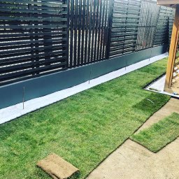 MEGIDO_Serwis - Układanie trawy z rolki przy ciemnym, nowoczesnym ogrodzeniu z poziomych desek i pergoli, widoczna warstwa białego podłoża pod trawnikiem i elementy drenażu.