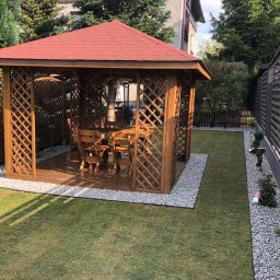 MEGIDO_Serwis - Drewniana altana z czerwonym dachem, otoczona zielonym trawnikiem i szarym żwirem, z widocznym stołem i krzesłami wewnątrz, częściowo zacieniona przez drzewa.