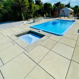 Nowoczesny basen z turkusową wodą i betonową obudową, z ukrytym zejściem, otoczony zielenią i placem zabaw w tle, pod błękitnym niebem.