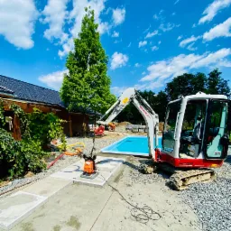 Mini koparka Takeuchi TB216 na żwirowym podłożu obok budowanego basenu z błękitną wodą, w tle budynek z ciemną dachówką i wysokie drzewo pod błękitnym niebem z białymi chmurami.