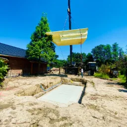 Kran opuszcza prefabrykowany basen do przygotowanego wykopu z betonową płytą fundamentową, widoczny budynek mieszkalny z ciemną dachówką i drzewo iglaste w tle.