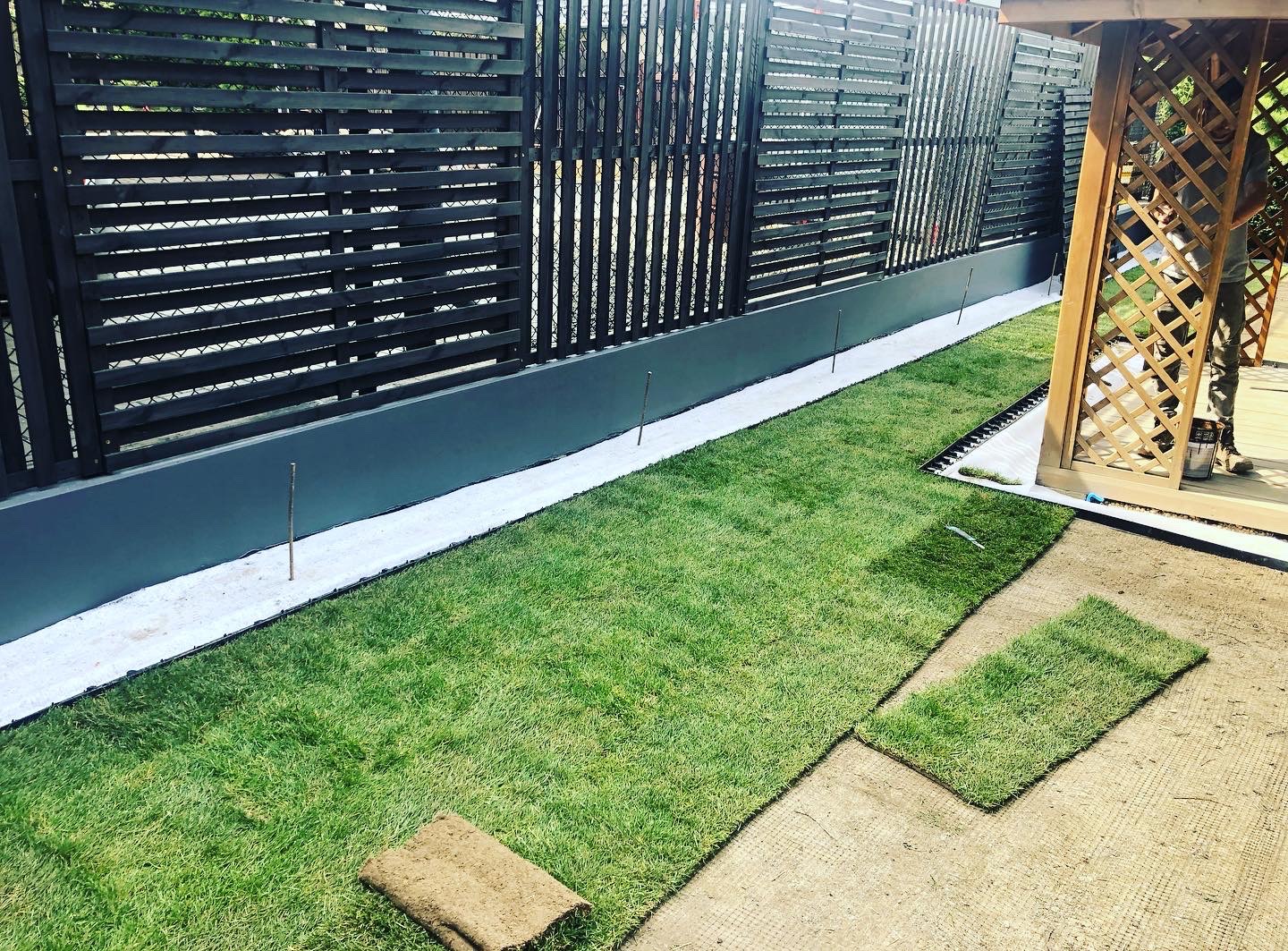 Układanie trawy z rolki przy ciemnym, nowoczesnym ogrodzeniu z poziomych desek i pergoli, widoczna warstwa białego podłoża pod trawnikiem i elementy drenażu.